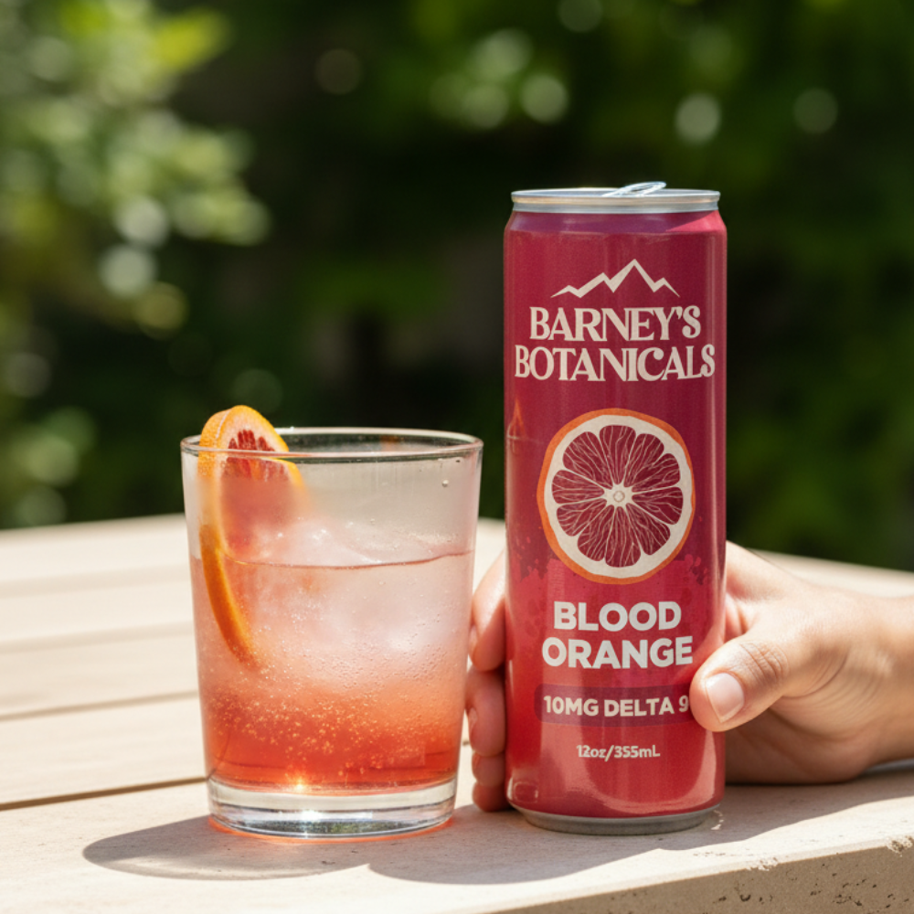 Hand holding a can of Barneys Botanicals Blood Orange drink next to a glass with a similar beverage outdoors.